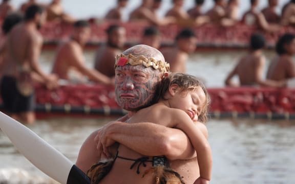 Wiremu Keretene (Ngāti Hine) holds a sleeping child during the waka display at Tii Beach this morning.