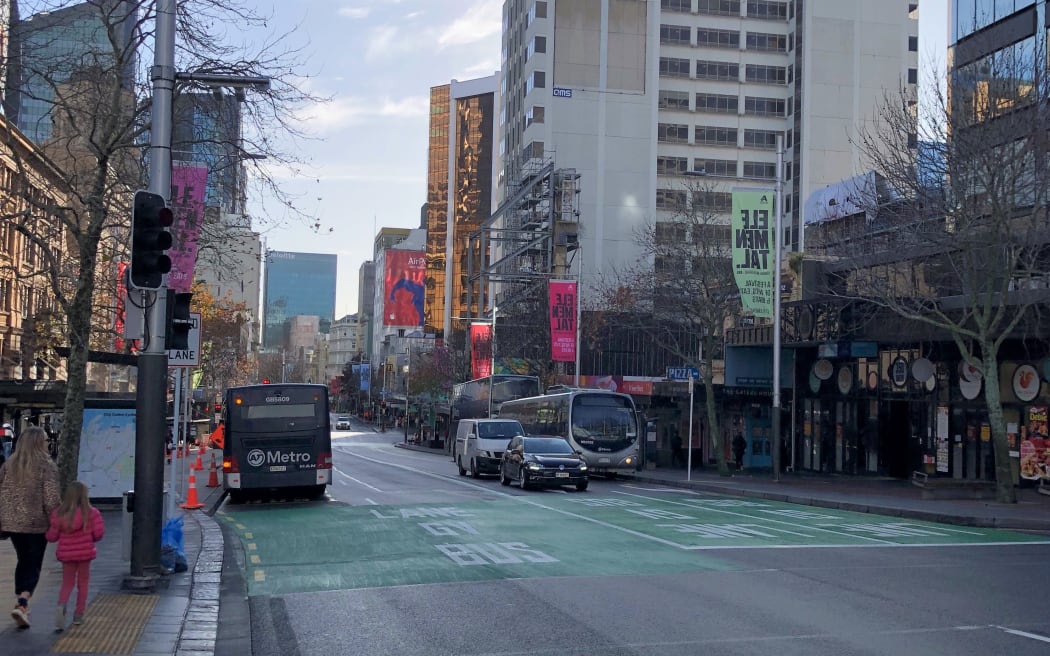 Queen Street on a quiet weekday afternoon. The sky is pale blue, the streets are in shadow. A few buses and cars busy the roads. A woman and her daughter walk along the sidewalk on the left.