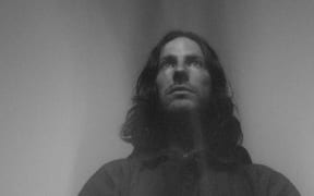 A monochrome photograph of a man looking into the middle distance. The camera looks up at his face.