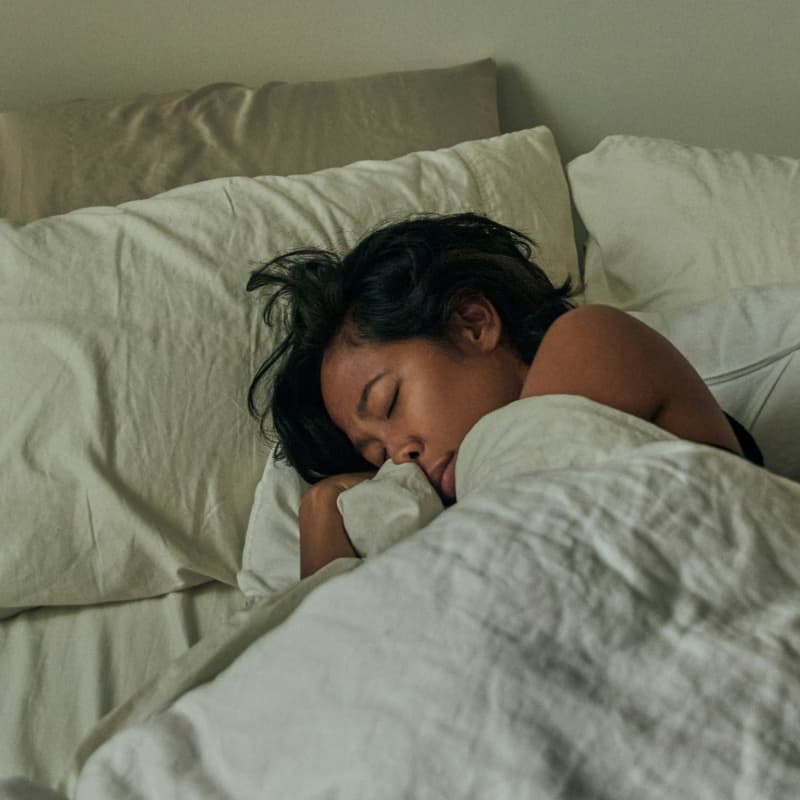 A woman with black hair curls up asleep in bed.