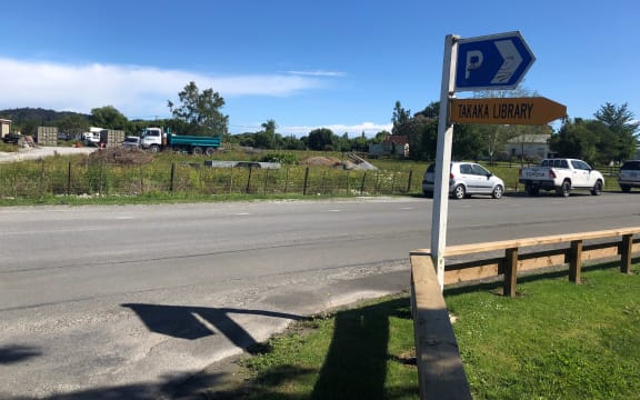 A photo of a country road taken from the roadside. On the far side is a lot with trucks and stacks of crates sitting idle. The grass is full of wildflowers. It is a sunny, blue sky day.