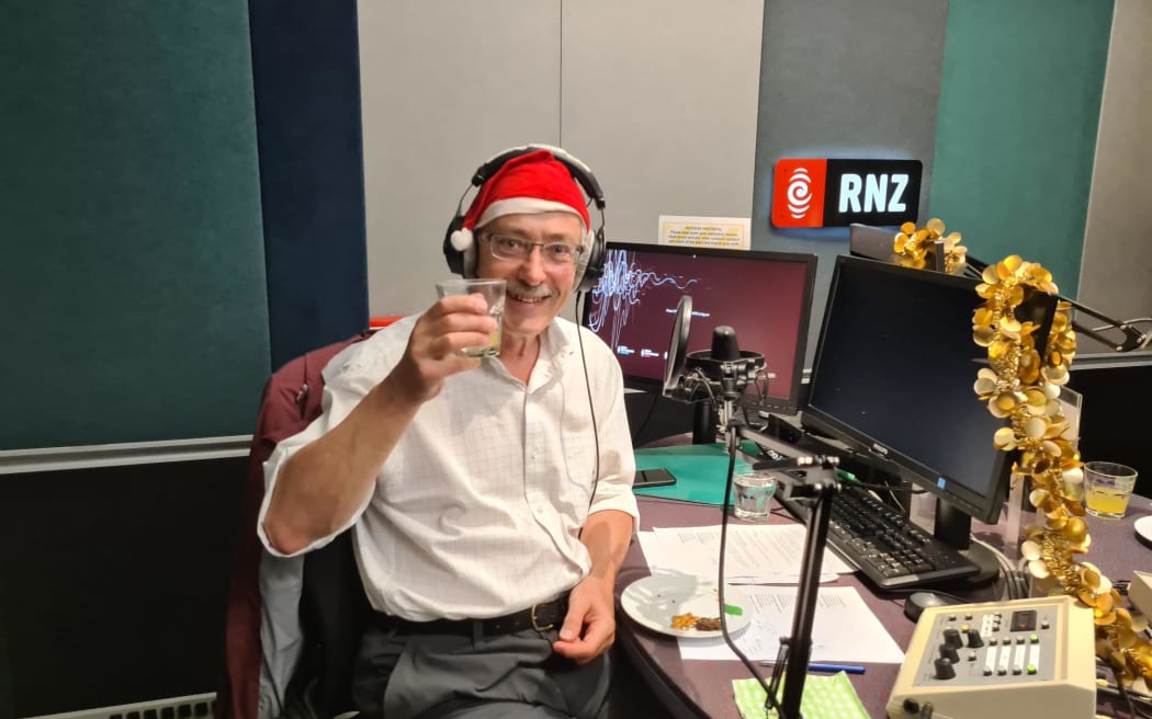 Rod Oram holding up a glass and wearing a Santa hat as he sits in a RNZ recording booth.