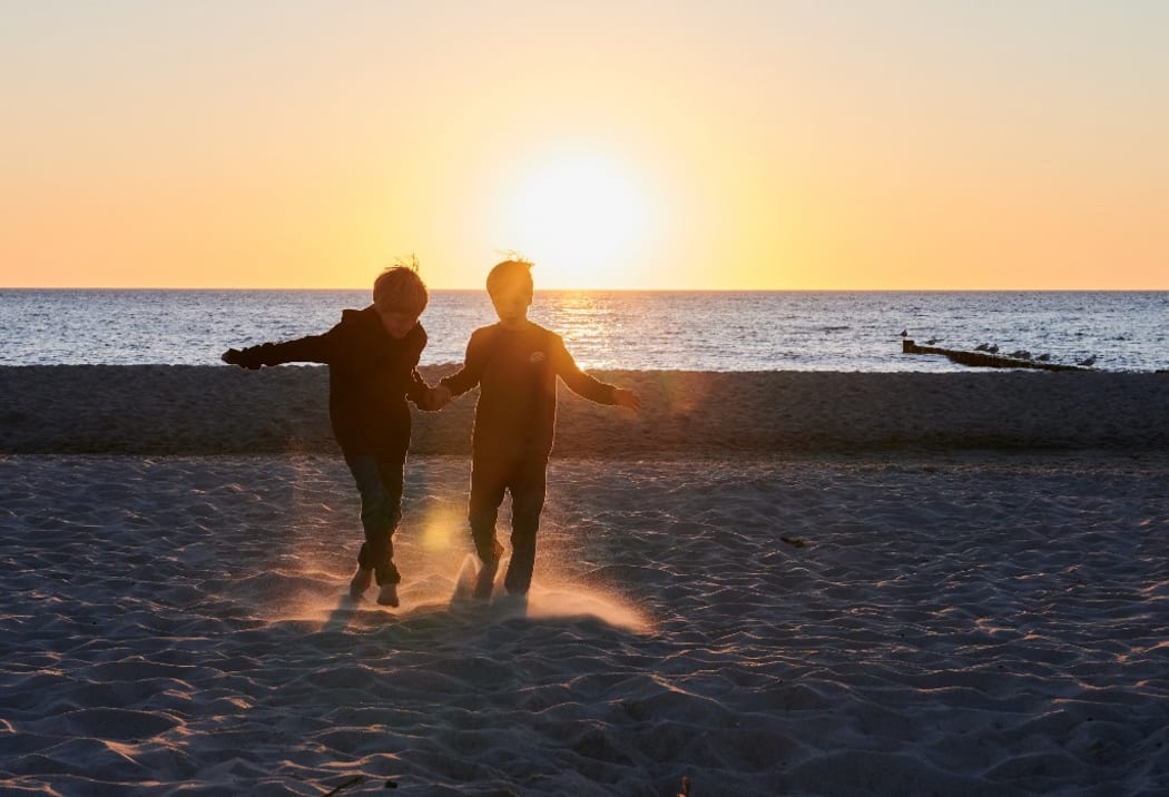 14 June 2020, Rostock: Two children walk on the sandy beach. The sun sets on the horizon of the Baltic Sea.