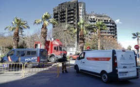 A police officer allows passage to a mortuary van at the multistorey residential block ravaged yesterday by a huge fire that killed at least four people, in Valencia on February 23, 2024. At least four people have died in a vast fire that ripped through a 14-storey apartment block in Valencia in eastern Spain, but officials warned today that the death toll could rise sharply. Fourteen people are still missing, the regional administrator said. (Photo by JOSE JORDAN / AFP)