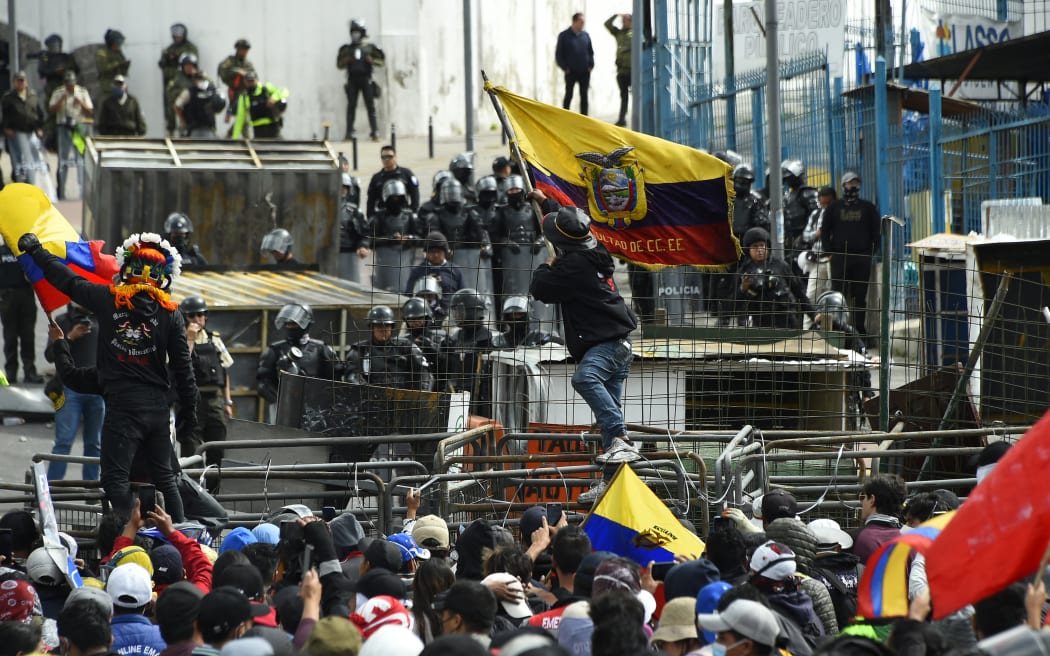 A demonstrator waves an Ecuadorean national flag while police officers stand guard in the surroundings of the National Assembly in Quito on June 25, 2022, when a parliamentary session called by opposition lawmakers for a no confidence vote against President Guillermo Lasso will be held in the framework of indigenous-led protests against the government. - An estimated 14,000 protesters are taking part in a nationwide show of discontent against rising hardship in an economy dealt a serious blow by the coronavirus pandemic. Protesters are demanding a cut in already subsidized fuel prices, which have risen sharply in recent months, as well as jobs, food price controls, and more public spending on healthcare and education. (Photo by Rodrigo BUENDIA / AFP)