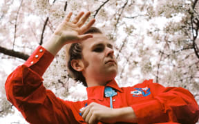 Ōtautahi/Christchurch based songwriter Sam Bambery wears a bright red Western shirt with slicked back hair under a cherry tree.
