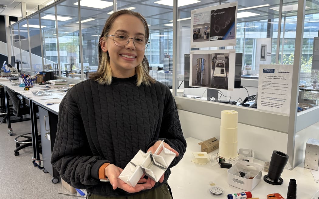 Research assistant Ella Fasciana holds up a small model of the SAR4SaR search and rescue device, made from tape and small pieces of corflute, while standing in a laboratory.