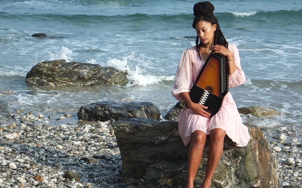 British folk musician Angeline Morrison sitting on a rock by the sea holding an autoharp.