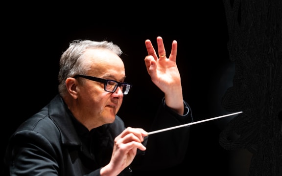 Conductor Marc Taddei holds a baton in one hand and raises the other as he conducts