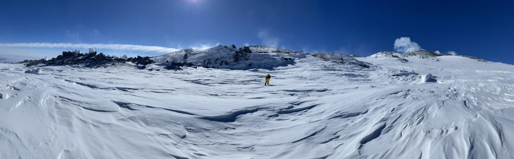 A panorama of ice and snow with dark volcanic rocky outcrops rising into a bright blue sky. A small figure can be seen in the middle of the panorama standing on the ice.