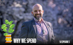 Smiling man in a patterned shirt and dark blazer stands outdoors in front of blurred purple foliage. He wears a pendant and a rainbow pin on his lapel. A cartoon plant growing out of stacked coins appears beside him, with bold white text reading 'Why We Spend' and smaller text in the corner: 'S4 Episode 5'