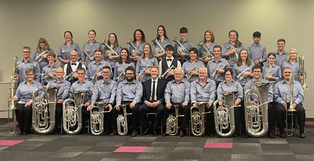The Rolleston Brass pose for a band photo, seated in rows inside a large room. They all wear blue shirts and hold their instruments, smiling.
