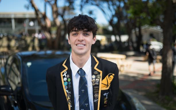 A portrait shot of Jesse outside in front of a car. He is wearing his school blazer which is black with yellow piping and has lots of badges on it.