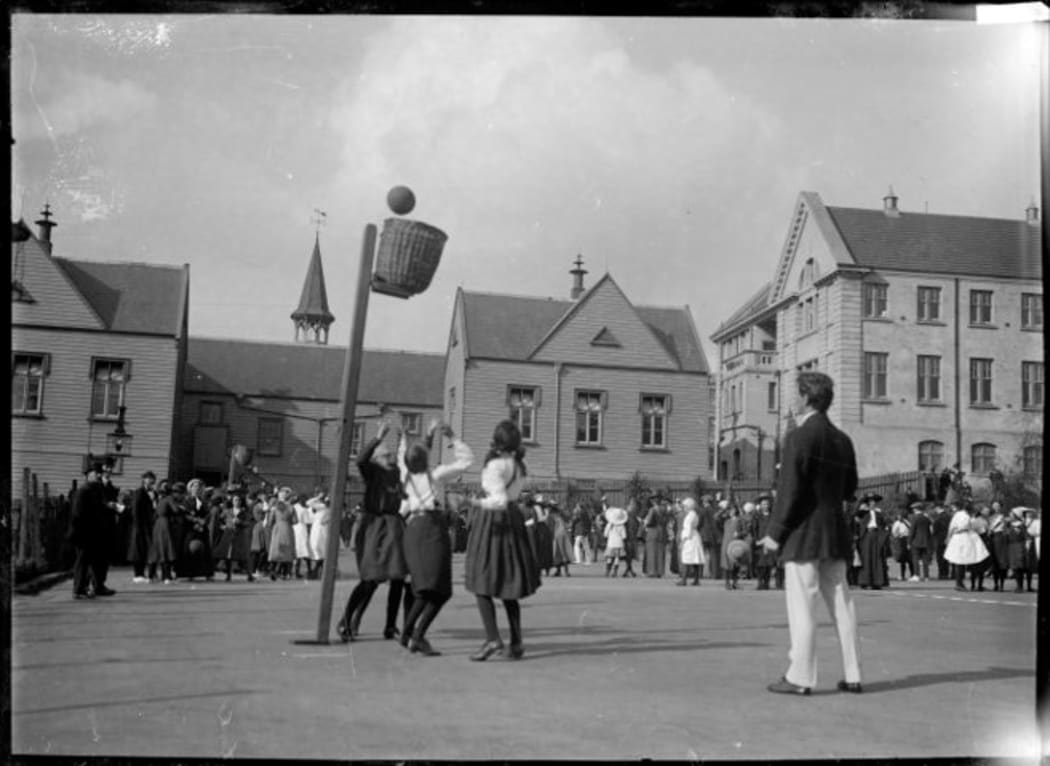 A game of basketball (later known as netball). Photograph shows a game in progress with the ball about to go into the basket (goal). In the background are spectators and unidentified school buildings. Taken by William A Price circa 1910.
