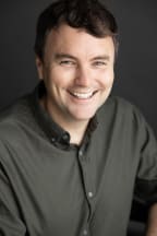 A headshot of Tom Sainsbury wearing a dark button up shirt and smiling nicely at the camera.