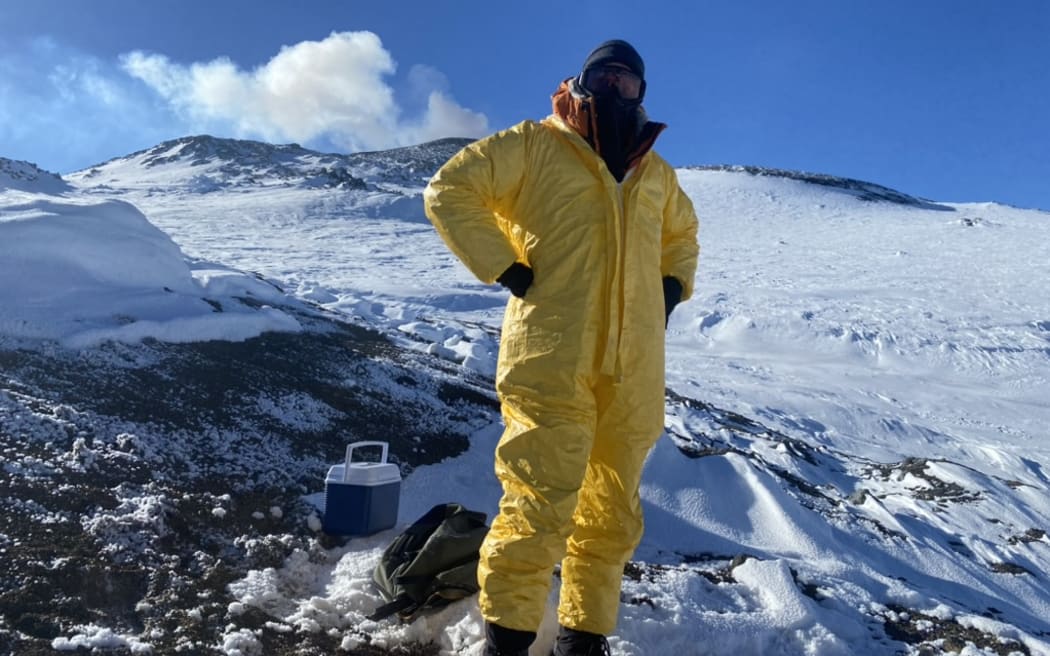 A person wearing a bright yellow hazmat suit, dark balaclava and eye goggles stands on a slope on dark volcanic rock and snow.