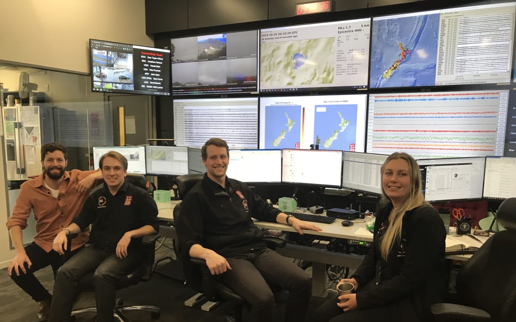 Four people on office chairs smile at the camera while sitting in front of an array of six computer monitors and ten large screens showing maps and data.