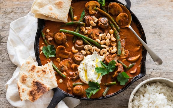A cast iron frying pan filled with a rich red-brown curry of mushrooms, cashews and green beans, garnished with flatbreads, coriander and yoghurt.
