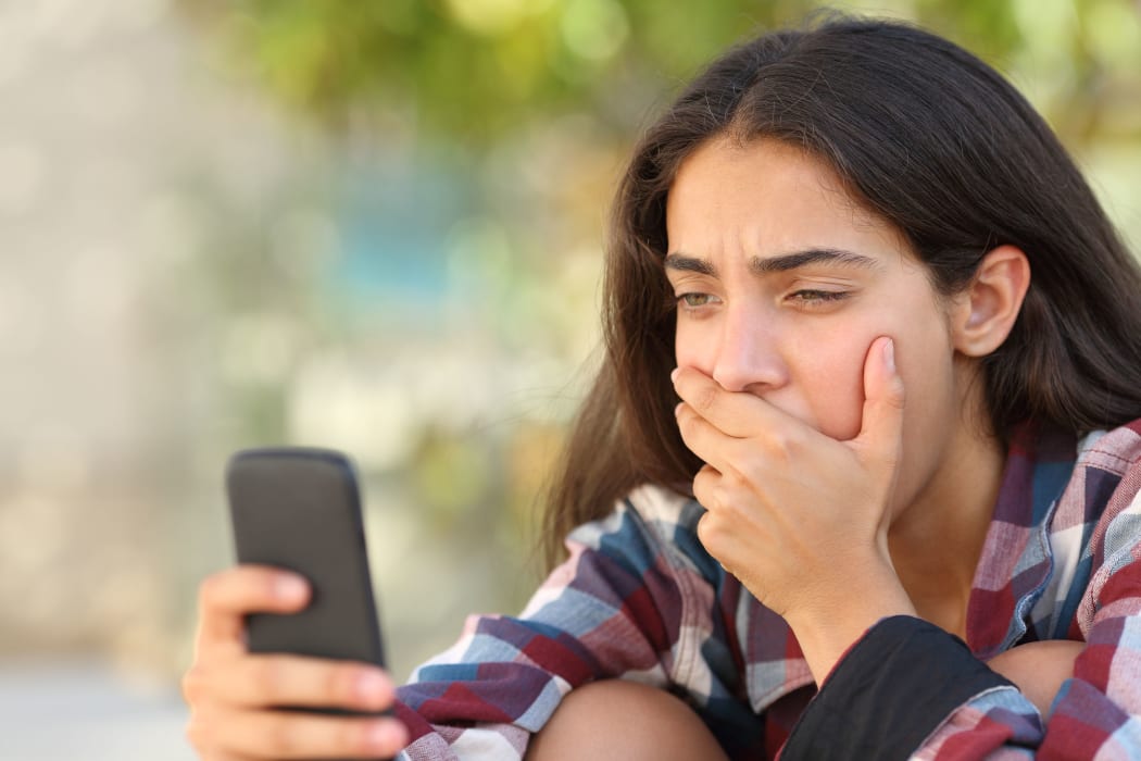 A photo of a worried teenager girl looking at her smart phone in a park with an unfocused background