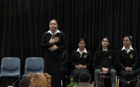 Tuhingaia is mid-speech, standing on a stage in school uniform holding her hands in front of her chest. Other students sitting on stage look on.