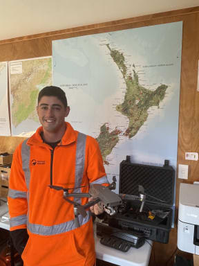 Jordan is wearing an orange high vis top in front of a map with New Zealand. He is holding a quad drone in his left hand, in front of the open padded box that it is stored in.