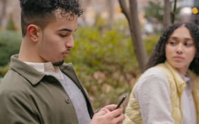 A young man looks at his phone while a young woman looks at him.