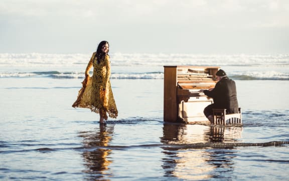 Music duo Allana Goldsmith and Mark Baynes are on a beach in the shallow. Allana holds her dress out of the water, Mark sits at an upright piano.