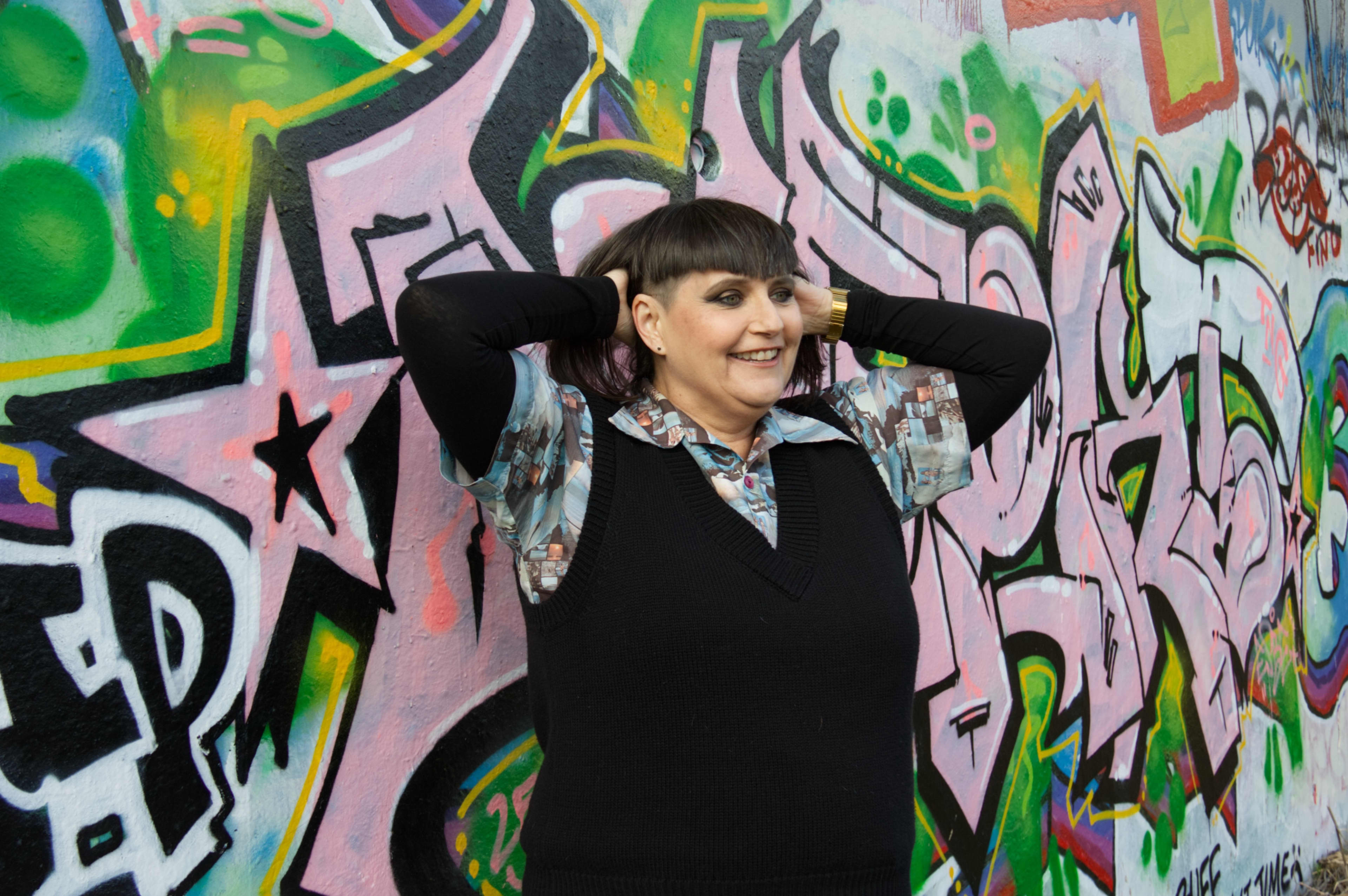 Author Pip Adam, wearing a black sleeveless vest over a colourful short sleeved shirt with black sleeves underneath, stands, smiling, in front of a wall of grafiti, raising her hands behind her head.