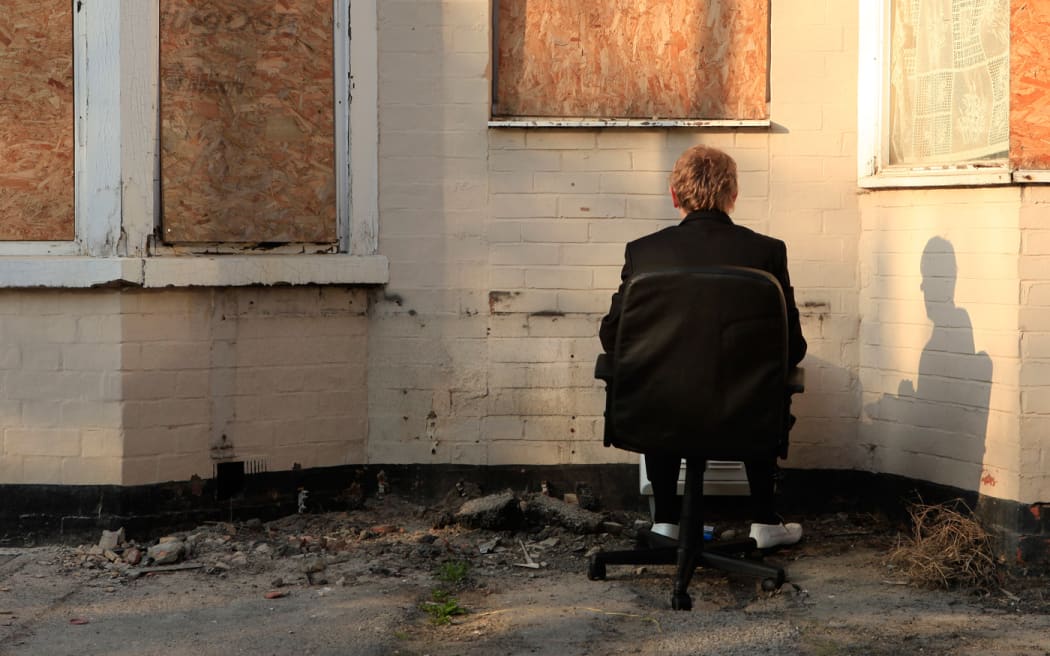 Man in chair sitting on street staring at a brick wall.