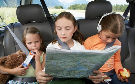 Three children in the back seat of a car, one drinking juice, another reading a print map, and the third with headphones and an apple.