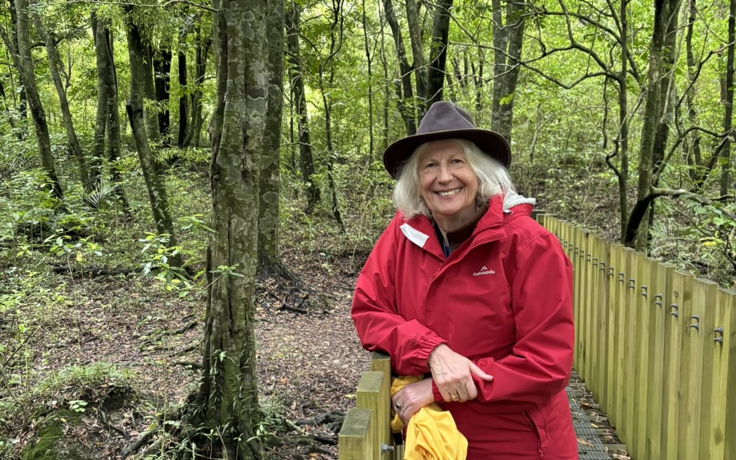 A woman with long grey hair under a wide-brimmed hat wearing a red rainjacket smiles while standing on a wooden bridge in lush green forest.