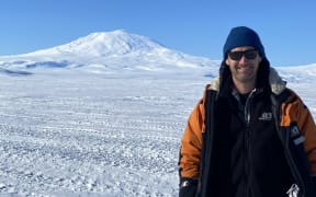 A man wearing a thick orange jacket, blue beanie and sunglasses stands smiling at the camera on an expanse of ice and snow criss-crossed with vehicle tracks. A snow-cloaked volcano is visible in the background, rising into a bluebird sky.