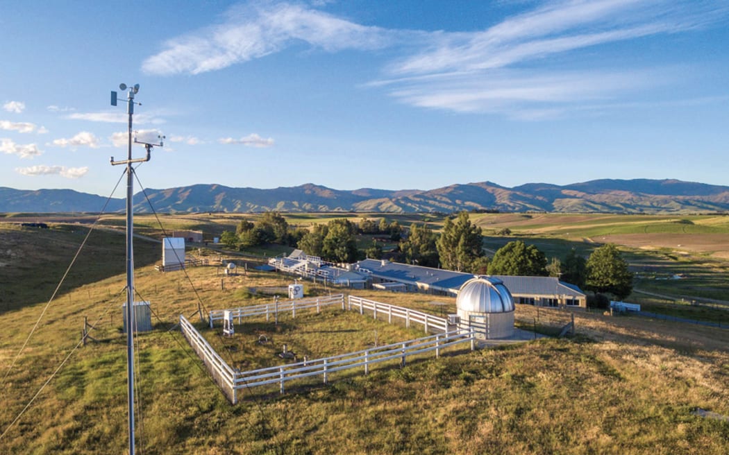 A wide photo of a cluster of buildings nestled behind trees on rolling green hills with mountains in the background beneath a blue, cloud-streaked sky. A silver metal dome is in the foreground next to a paddock enclosed with a white wooden fence.