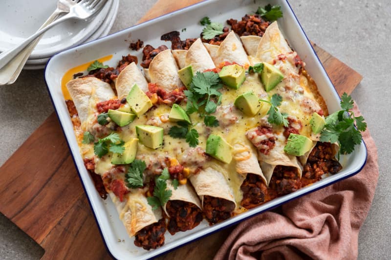 A rectangular dish of tofu and black bean enchiladas topped with avocado and coriander leaves.