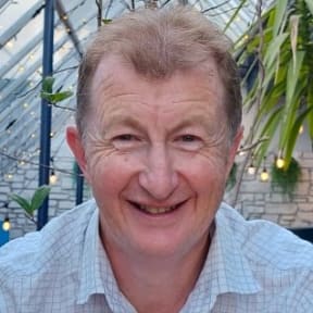 Dr Brent Gilpin smiles at the camera. He is wearing a white shirt. In the background are hanging lights and plants.
