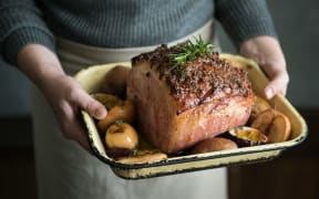 A person wearing an apron holds a casserole of baked ham with roasted apples on the side.