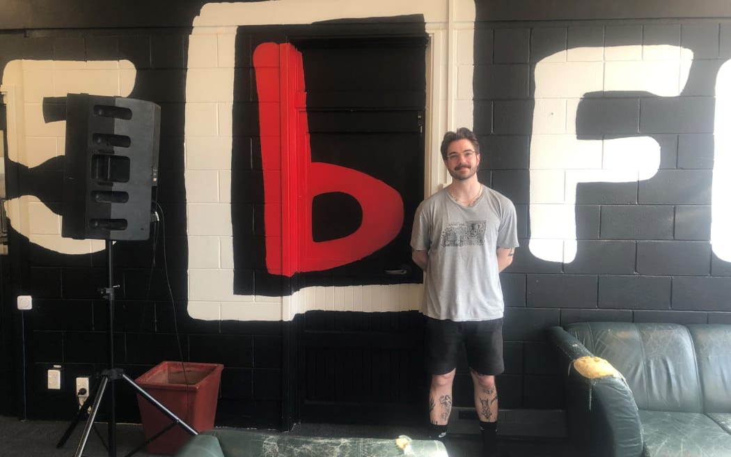 Tom stands in front of a black wall emblazoned with a large "95bFM" logo. To his right is a speaker on a stand. In front of him are a pair of worn-down green leather couches.