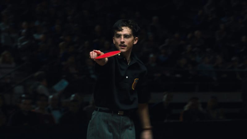 Timothee Chalamet, a young man with a moustache and glasses, holds a red ping-pong racquet as he points a finger.