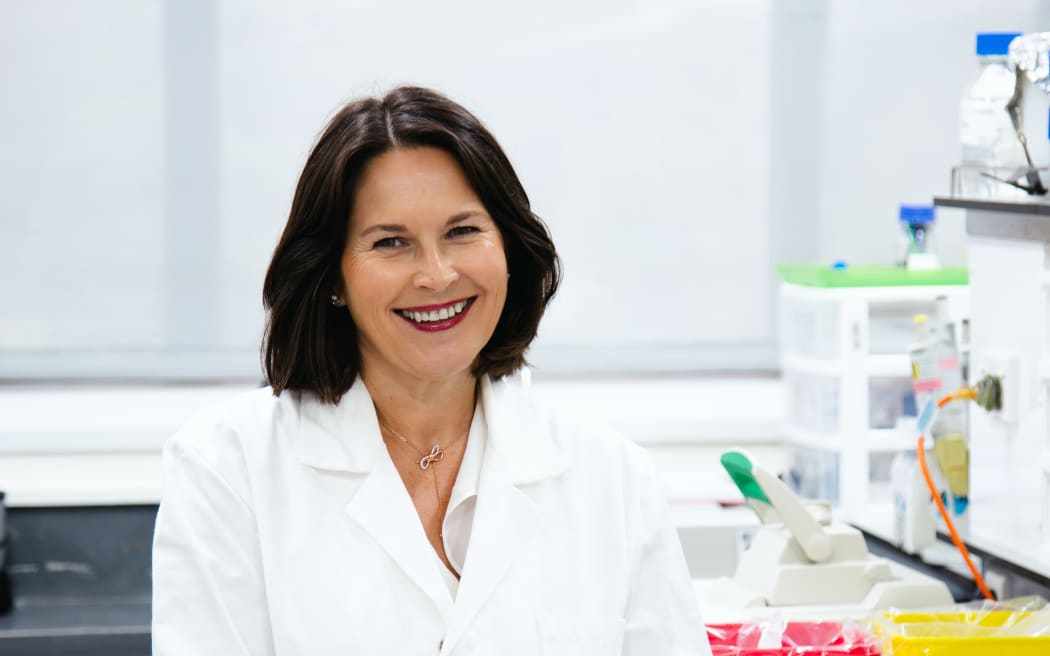 A headshot of a smiling woman with shoulder length brown hair in a lab coat at a white lab bench.