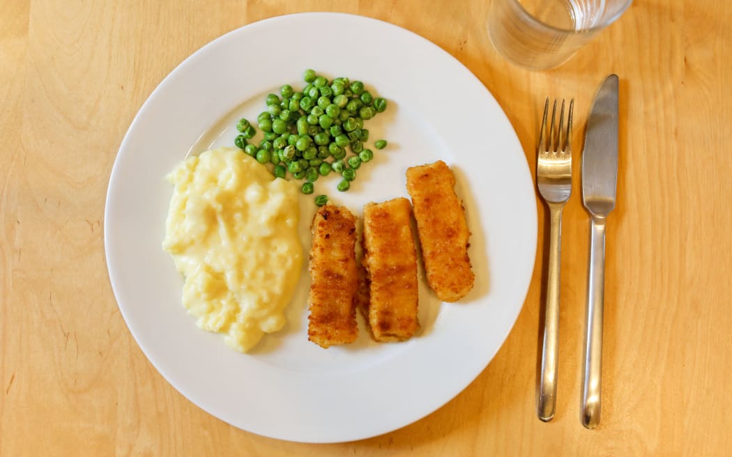 A plate with three fish fingers, mashed potatoes and peas stands on a table.