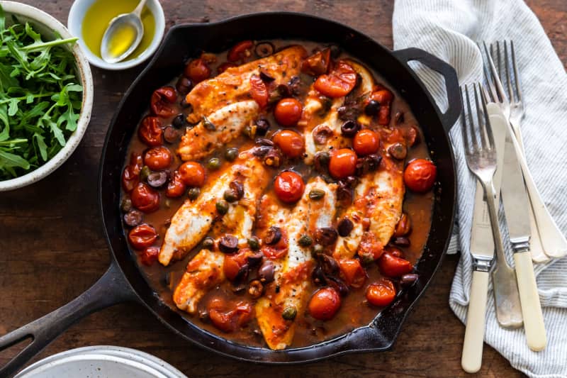 A round cast iron frying pan on a wooden board containing a dish of white fish fillets cooked in a sauce of tomatoes, capers and olives.