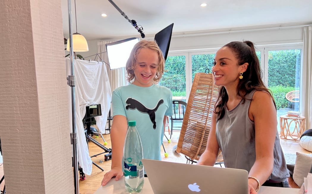 Sonia Gray and a child sit at a table, smiling. Sonia is holding an Apple MacBook. In the background is a makeshift film set, lights and a white sheet draped as a backdrop for a camera shot.