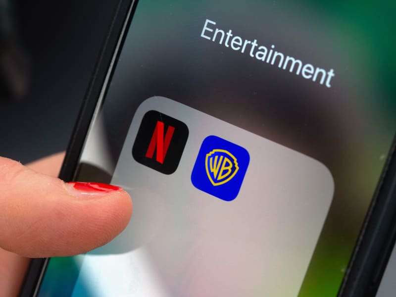 A finger taps an app folder field on a smartphone with the logos of the streaming platform Netflix (L) and the film studio Warner Brothers.