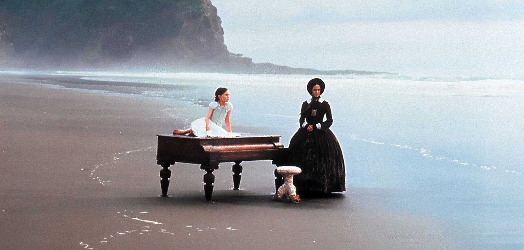 A grand piano sits on a wind-swept beach, a child sits on top and a black-clad woman stands beside them.