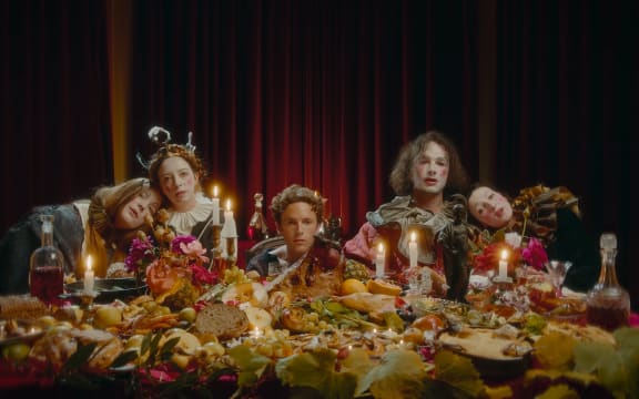 Still from the video to Soaked Oats' song 'Something'. The band and friends seated behind a sumptuous feast of food.