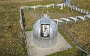 An aerial photo looking down at a metal dome on a concrete pad. A hatch on the dome's roof is open, revealing a machine inside.