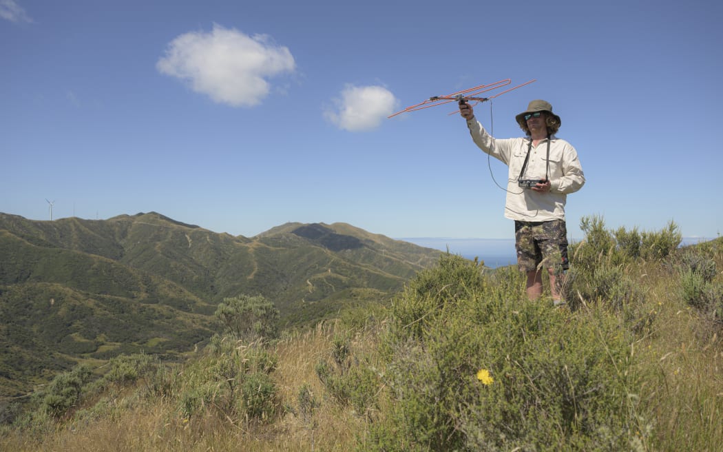 A man in a wide-brimmed sunhat standing on top of a grassy hill holding up a large antenna