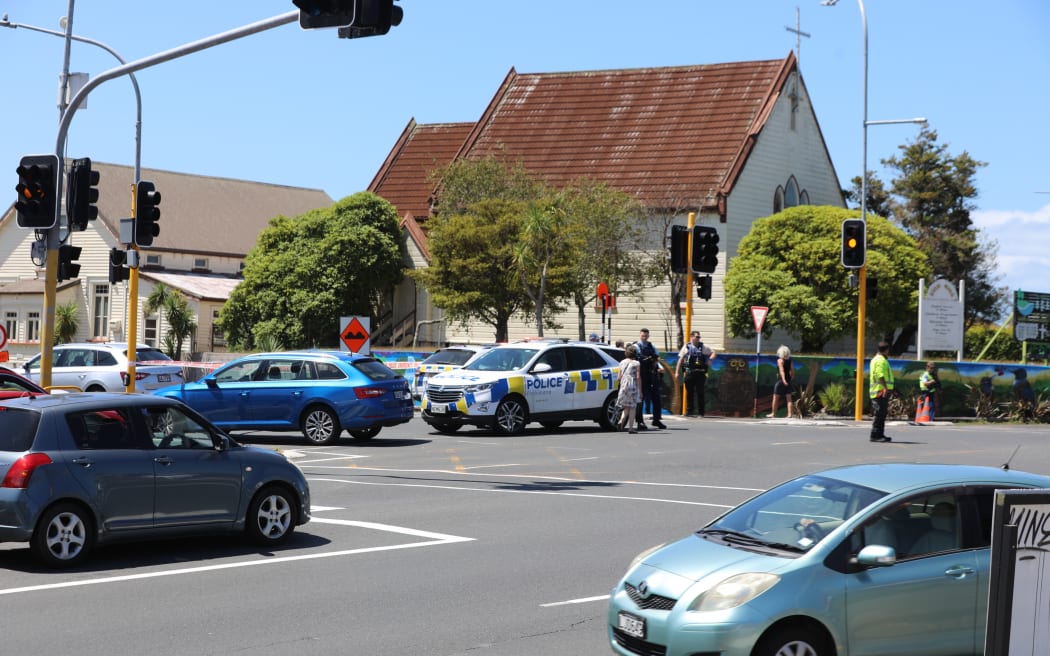 Lights are out at Birkenhead Ave intersection with Onewa Road and traffic is being manually directed by a person in a high-vis vest while armed police guard a cordon.