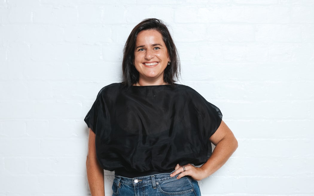 Sophie is standing against an off white background. It shows her from the waist up. She is wearing blue jeans and a black top tucked in at the waist. She had her left hand on her hip in a kind of classic 90s pose.