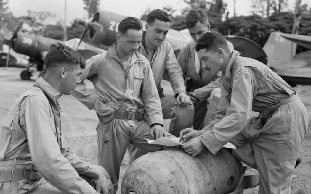 Group of pilots gathered around a bomb on a trolley to discuss air strike. Unknown location. 
Corsairs in the background believed to be on charge to No. 31 Servicing Unit.